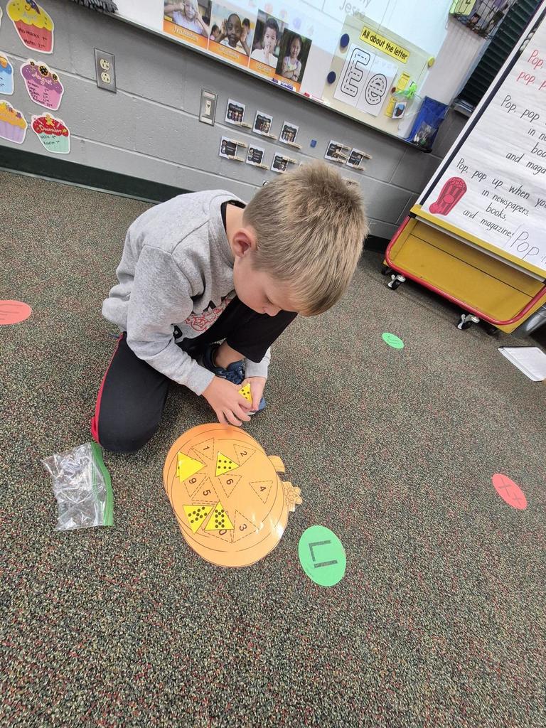 A child kneels on the carpet arranging yellow triangles on an orange pumpkin-shaped number-matching activity mat.