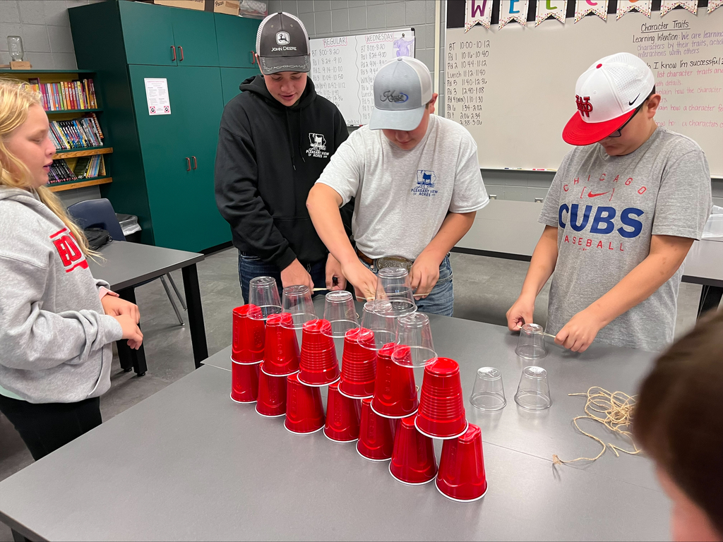 Four middle school students work together at a table stacking red and clear plastic cups during a teamwork activity using string and rubber bands.