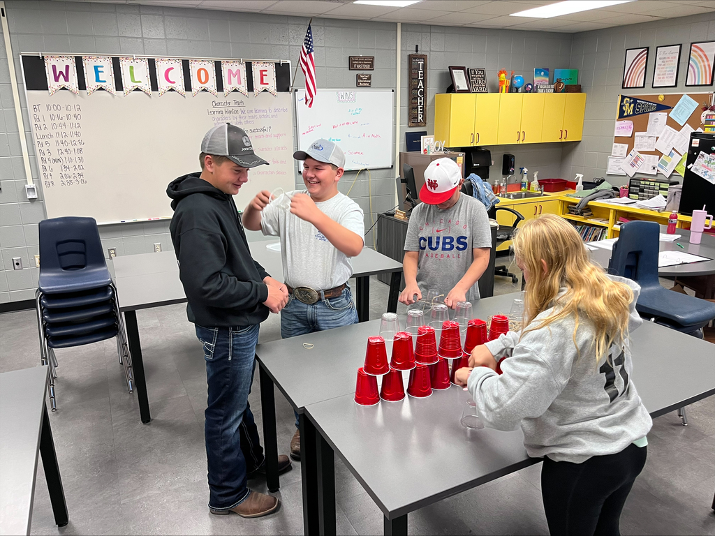 Students collaborate in a classroom on a cup-stacking challenge, smiling as they use teamwork and problem-solving skills.