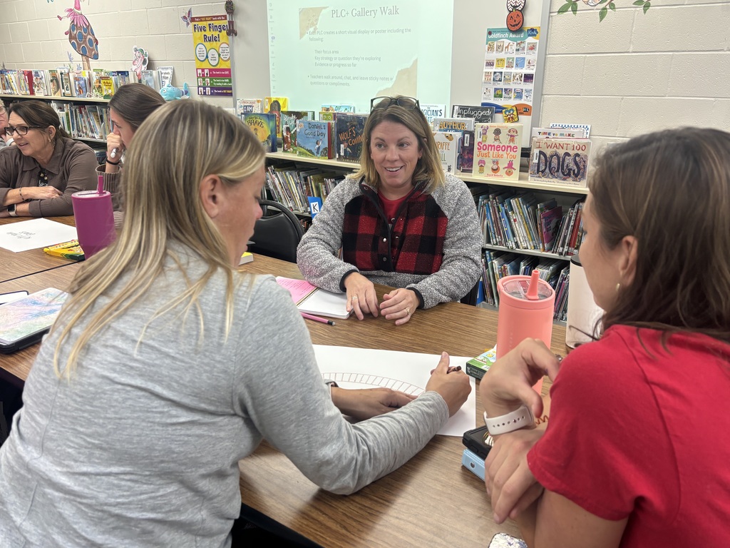 Teachers sit together at a table in a school library during a professional learning session. One teacher in a red and black plaid vest speaks as others listen and take notes.