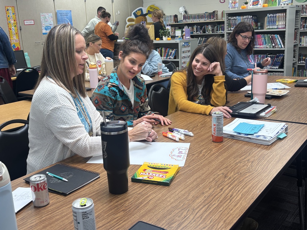 A small group of teachers gathers around a library table, sketching ideas on poster paper with colorful markers. One teacher writes while the others discuss and smile.