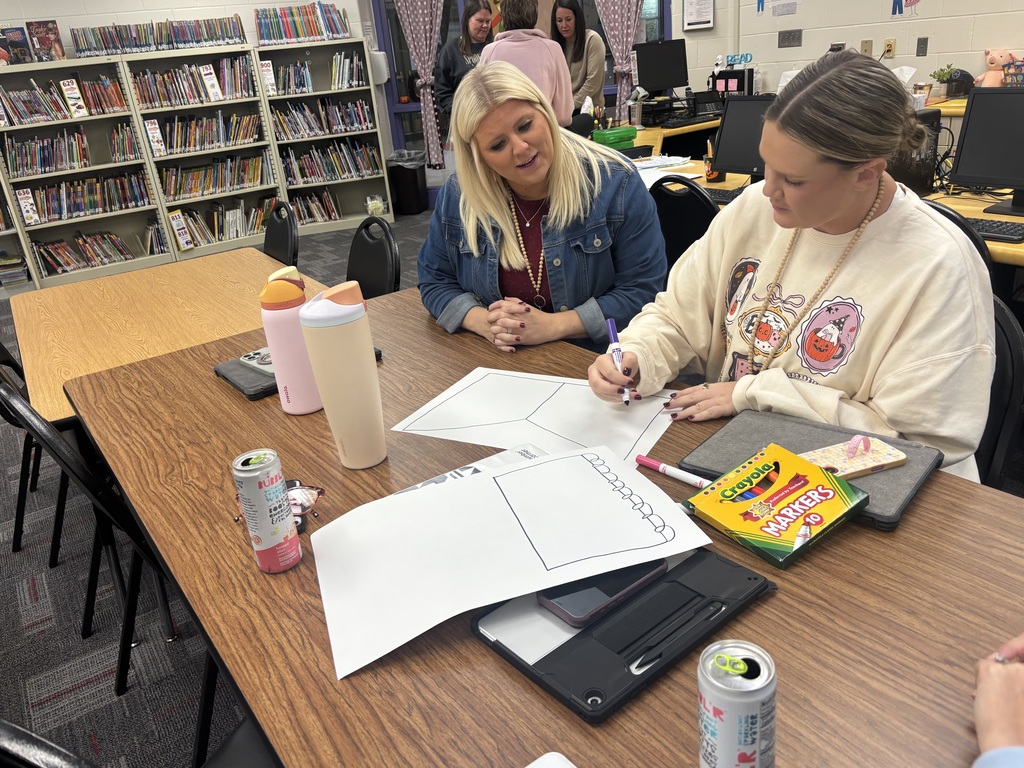 Two educators collaborate at a library table, one drawing on a large sheet of paper while the other looks on and smiles.