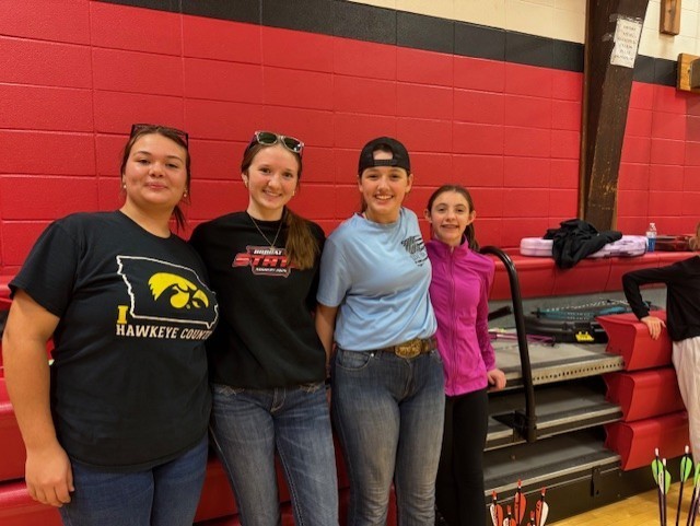 Four students stand in front of red gym bleachers smiling for the camera. They wear casual clothes and T-shirts representing different schools and programs.