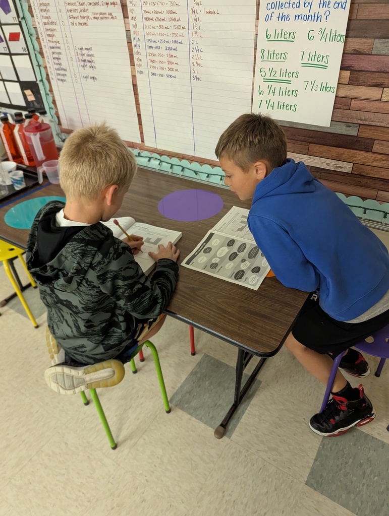 Two students sit together at a classroom table completing math problems in a workbook.