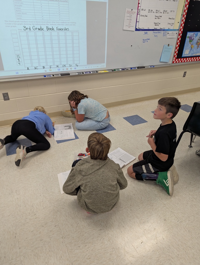 Four students work on math activities in front of a whiteboard displaying a chart titled “3rd Grade Book Favorites.”