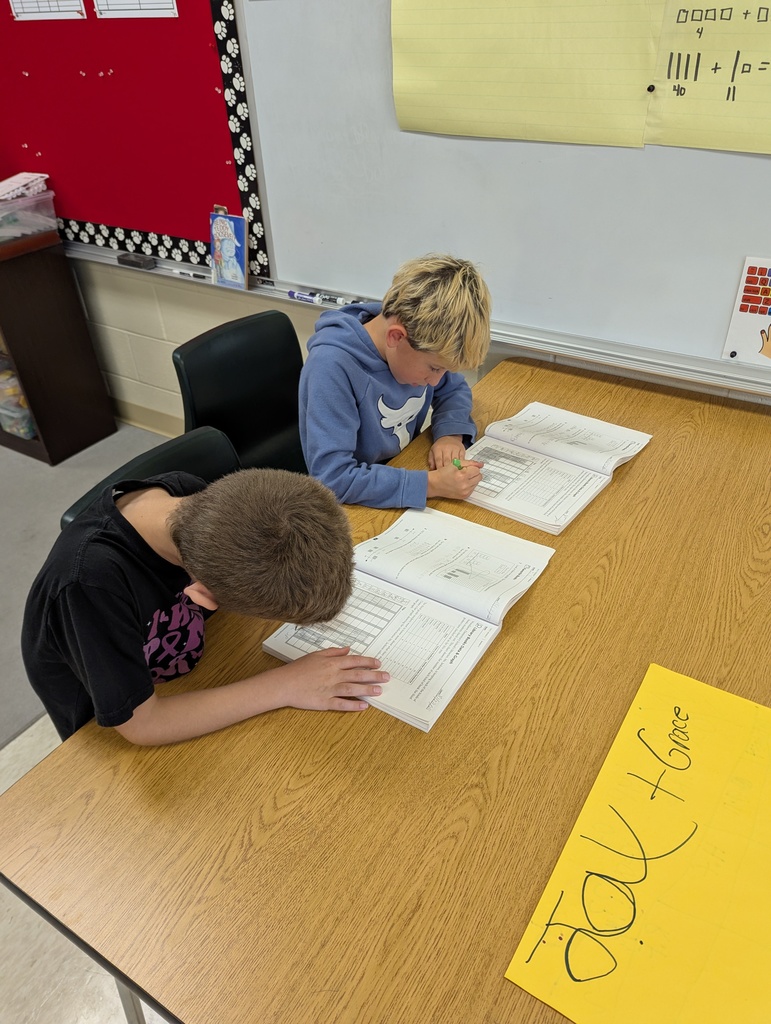 Two students sit at a classroom table working in math workbooks.
