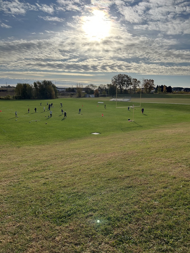 Students play soccer on a large grassy field under a bright morning sun.