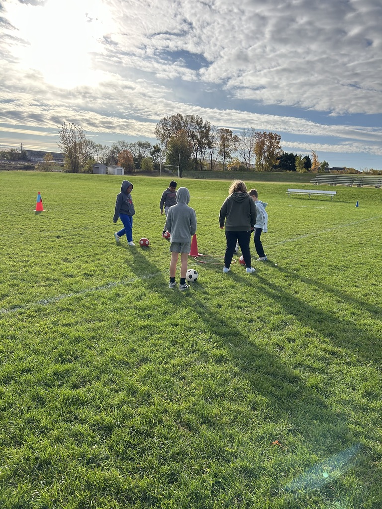 A small group of students practice soccer skills near orange cones on a sunlit grassy field.