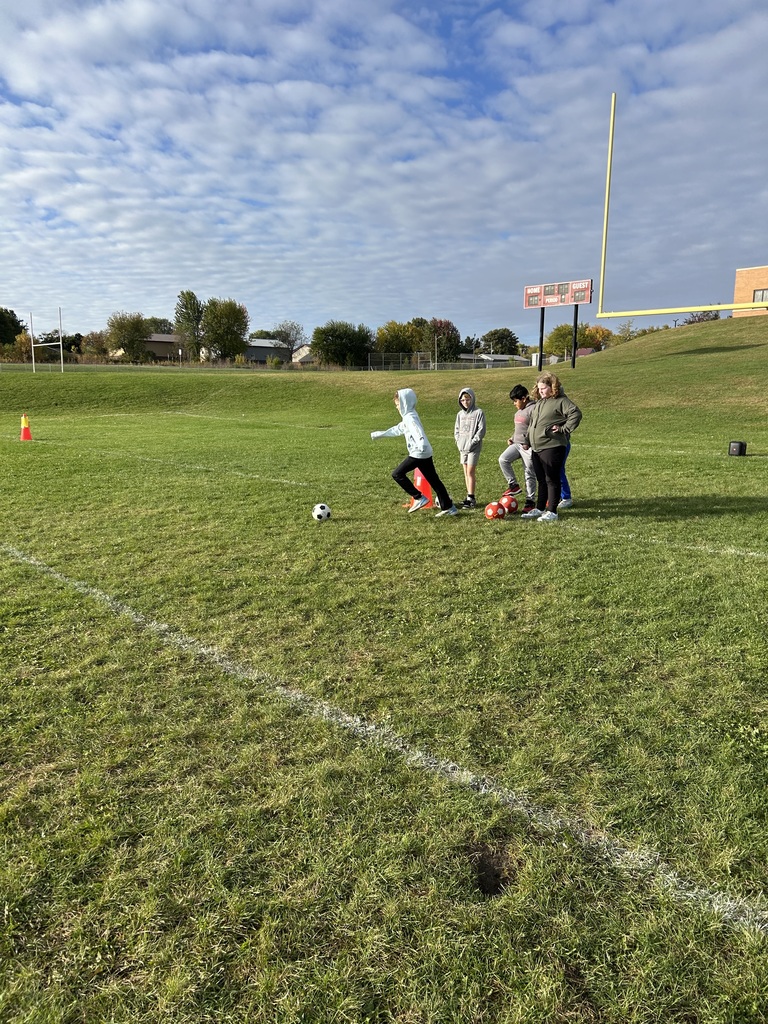 A student kicks a soccer ball while classmates watch nearby on a green field.