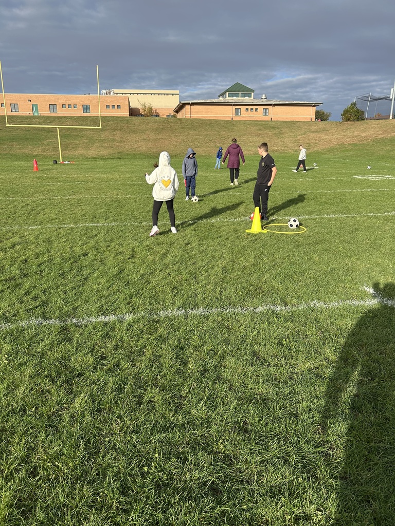 Students wearing hoodies and jackets practice soccer drills on a school field.