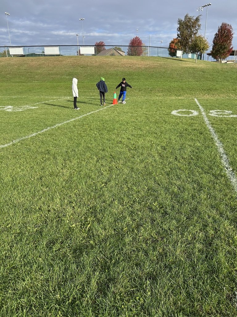 Three students stand near orange cones as one prepares to kick a soccer ball on a field marked with yard lines.