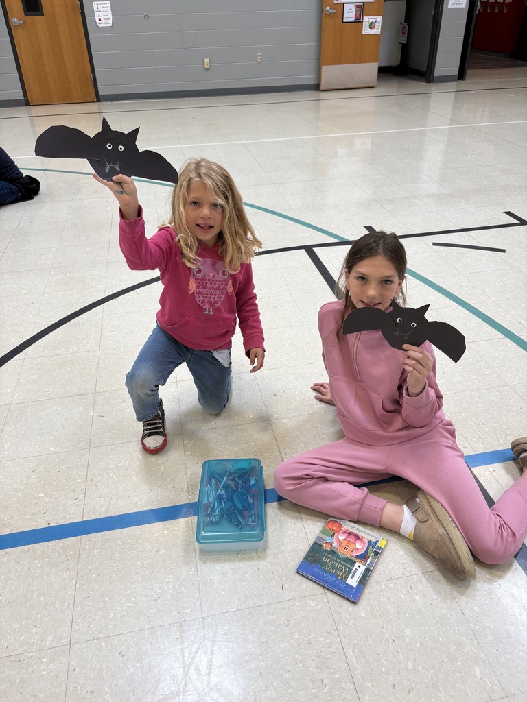 Two young students sit on a gym floor holding up black paper bats they made during a craft activity. One student has a book and supply box beside her.