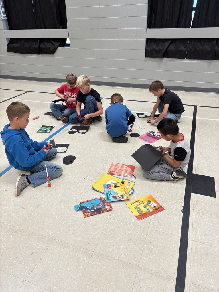 A group of children sit together on the gym floor cutting and gluing pieces of black paper to create bat crafts.