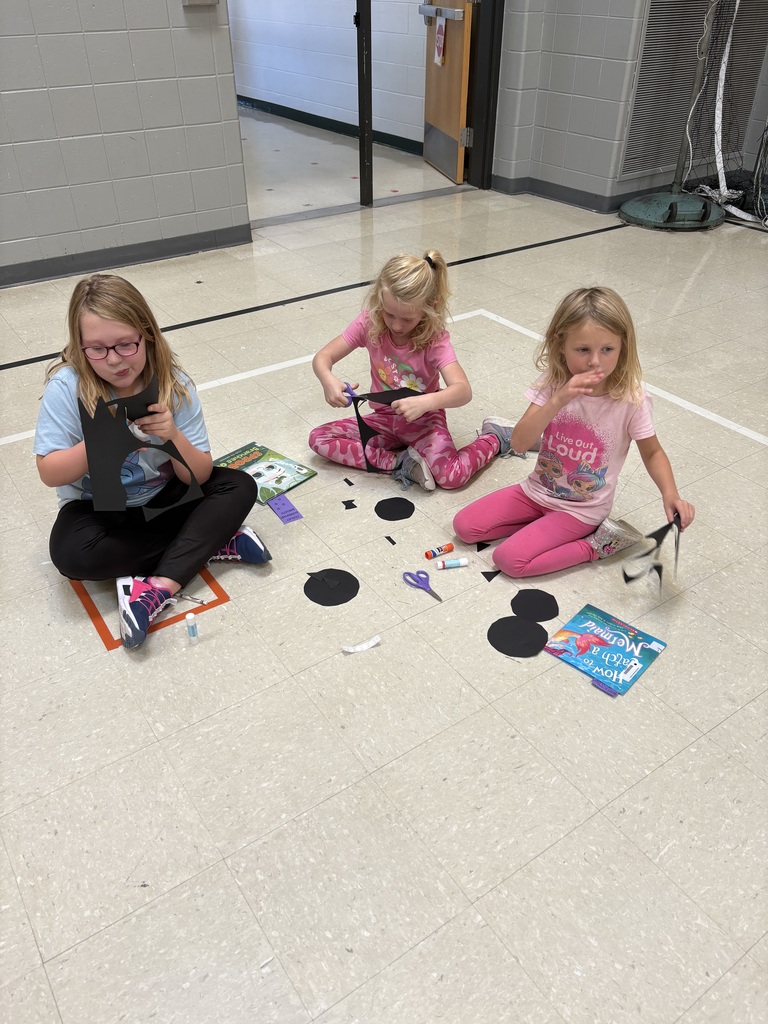 Three young students sit on the gym floor cutting shapes from black paper for bat crafts.