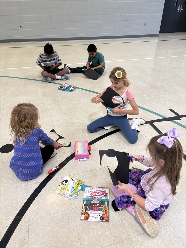 Several children sit in small groups on a gym floor working on Halloween bat crafts.