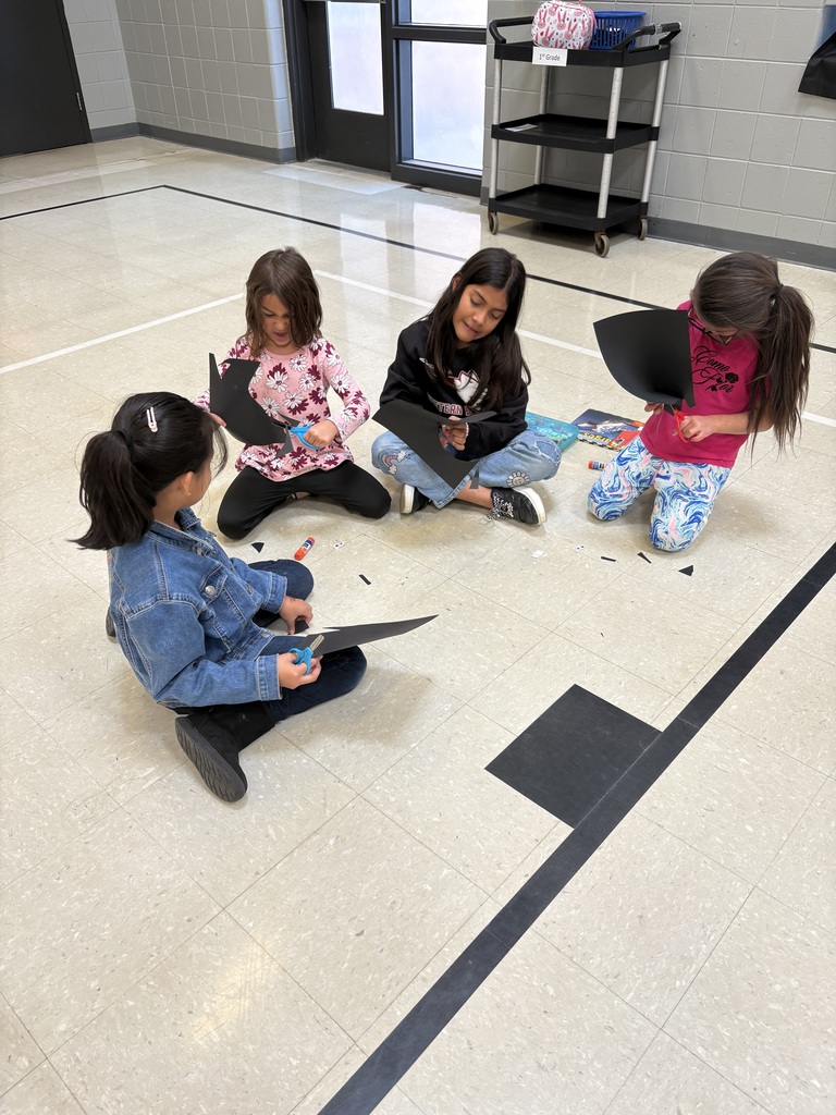Four children sit on the gym floor cutting black paper and gluing pieces together to make bat crafts.