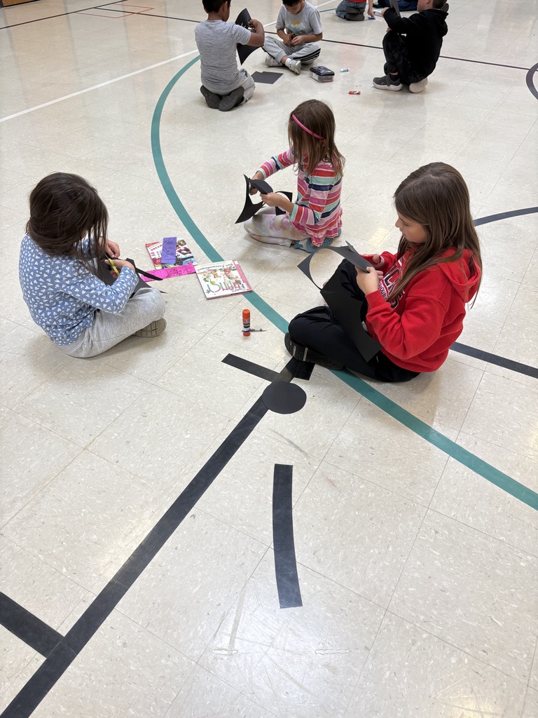 Students sit in pairs on a gym floor, cutting out shapes from black paper as part of a craft project.