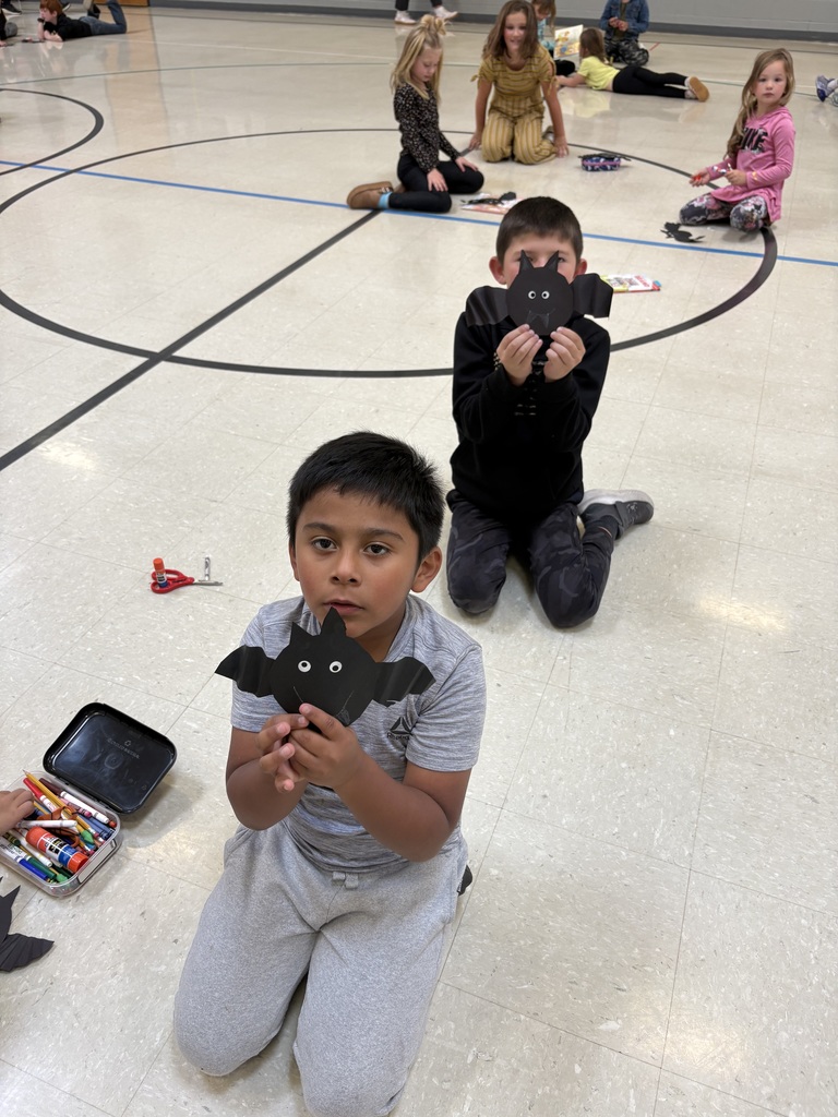 Students kneel on the gym floor showing their completed black paper bat crafts.