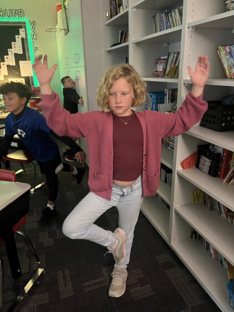 A student stands in a classroom balancing on one foot in a yoga pose, arms raised and eyes closed.