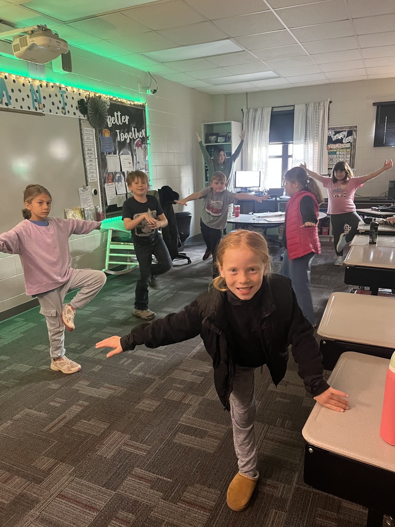 A group of elementary students in a classroom stand in yoga poses, balancing on one leg with arms outstretched.
