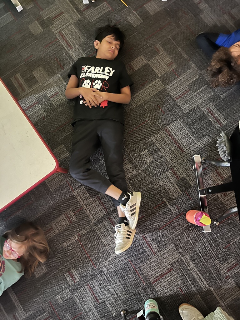 A student wearing a black “Farley Elementary” shirt lies on the classroom carpet with eyes closed and hands resting on their stomach, participating in a mindfulness or relaxation activity.