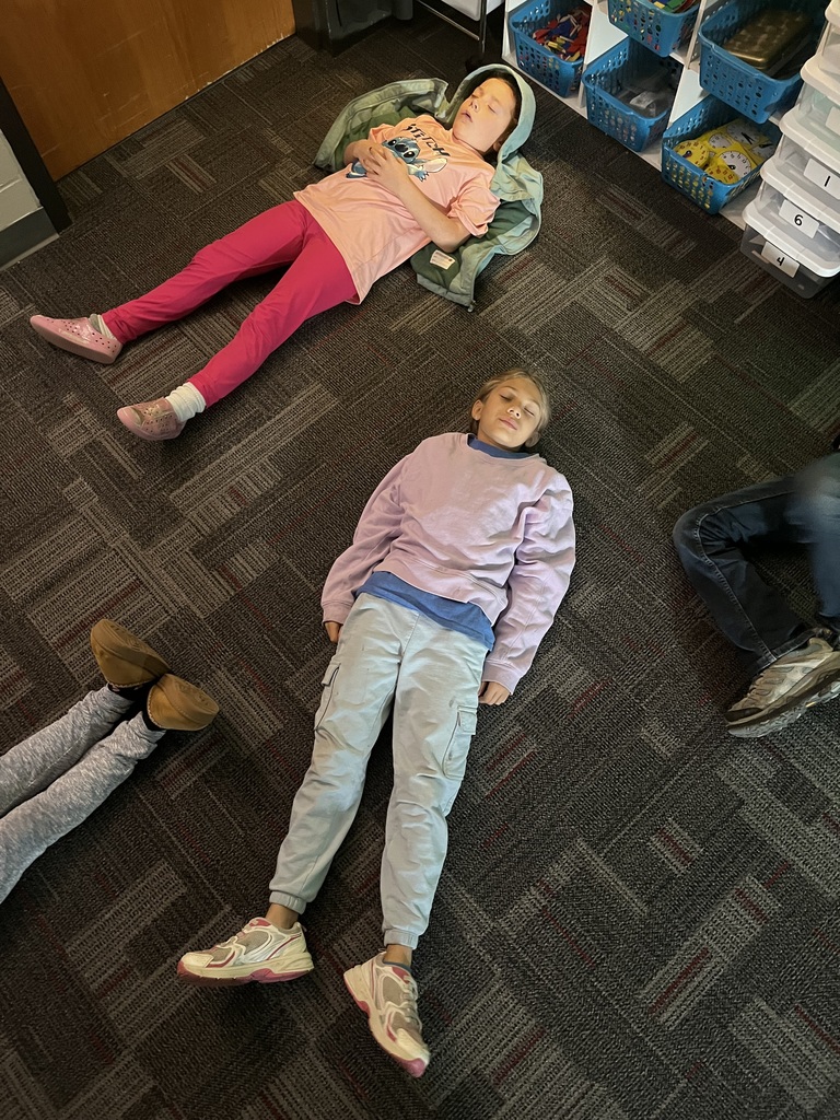 Two students lie on the classroom floor with eyes closed, relaxing quietly during a calm mindfulness activity.