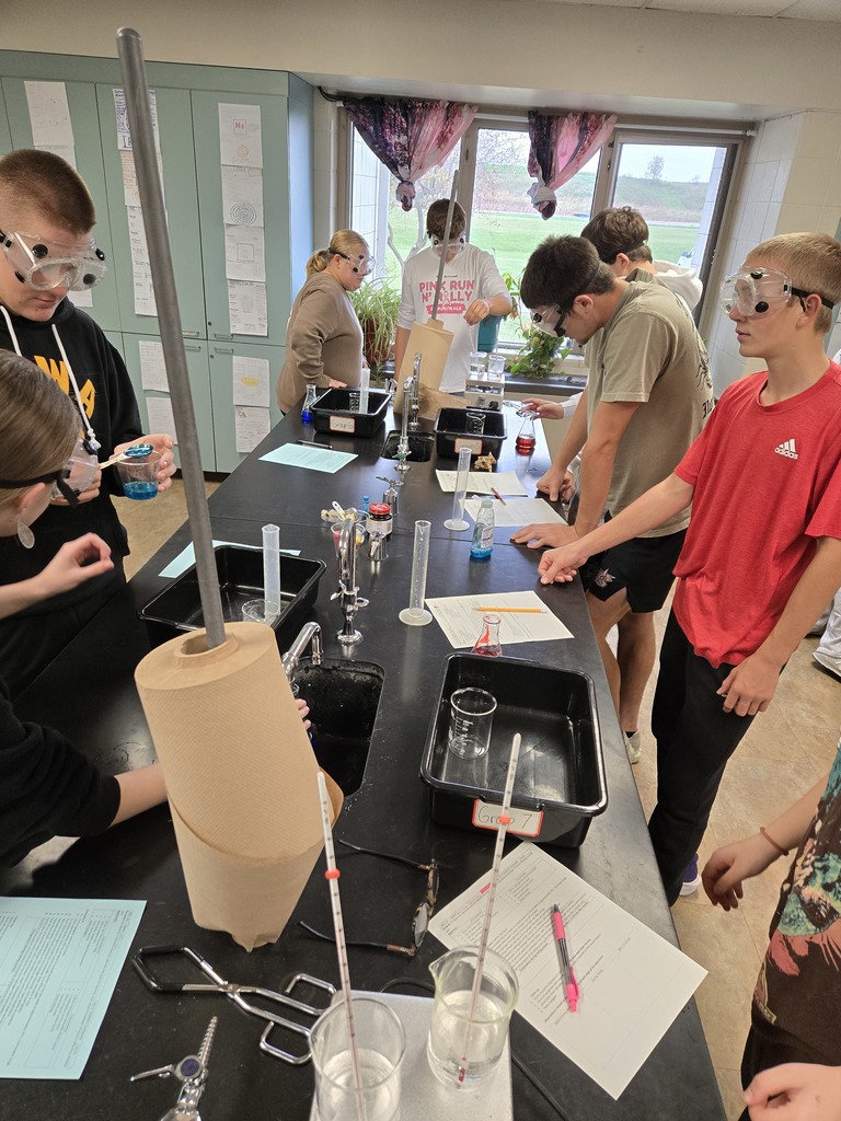Students wearing goggles stand around a lab table mixing colorful liquids in beakers and flasks.