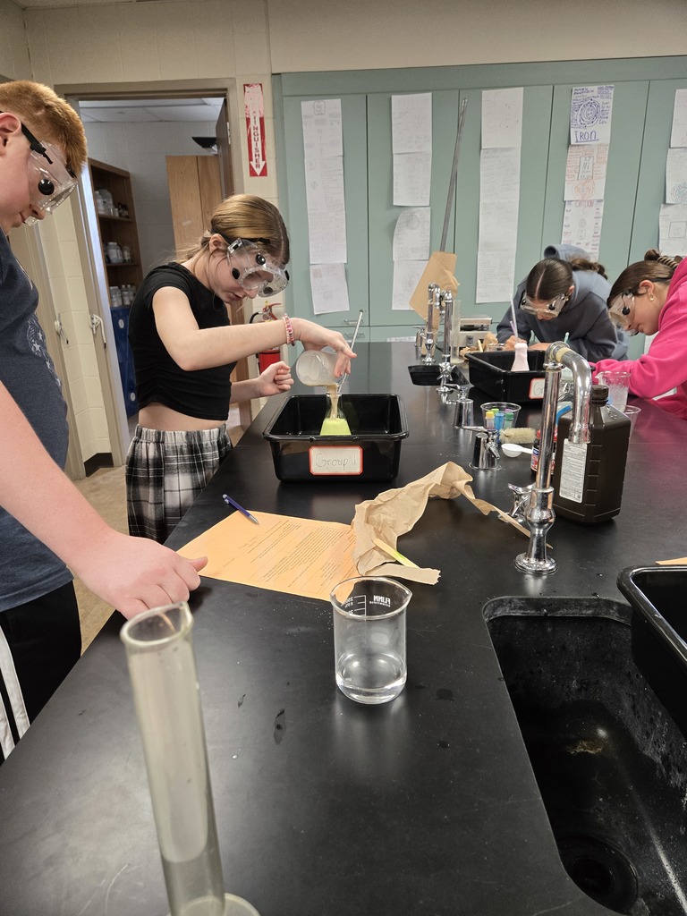 Students in safety goggles pour liquid from a beaker into a flask labeled “Group 8” during a classroom chemistry experiment.