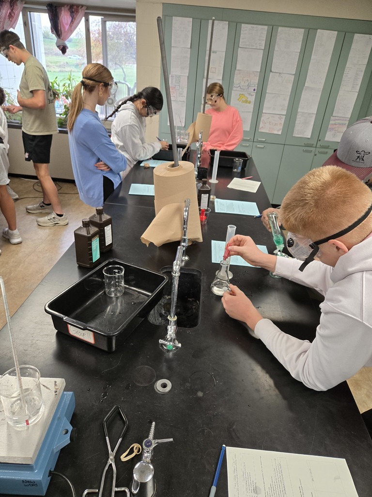Students in goggles perform a hands-on experiment at lab tables, mixing clear and colored liquids in flasks and beakers.