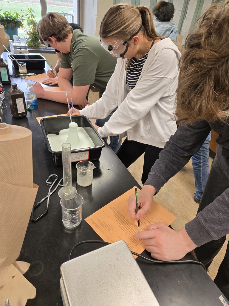 Students in goggles record observations on orange worksheets while watching a foamy reaction take place in a flask.