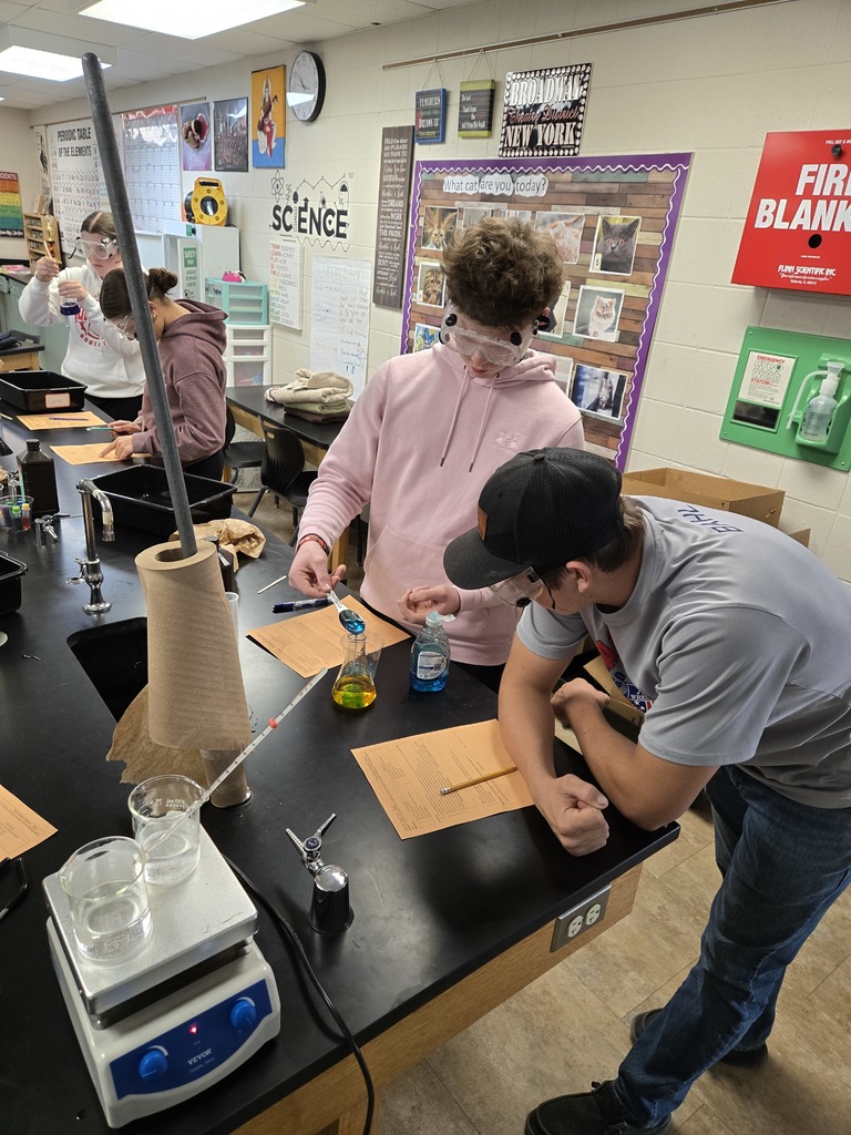 Students wearing safety goggles work together on a chemistry lab at a black science table. One student in a pink hoodie pours liquid into a beaker while another leans in to discuss the lab sheet.
