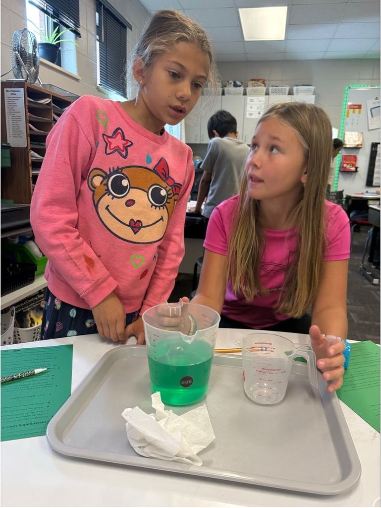 Two students collaborate on an experiment involving green liquid in a measuring cup.
