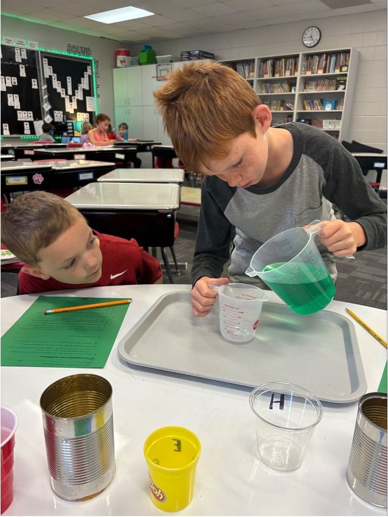 Two students work together at a table measuring green liquid into a smaller cup using clear plastic measuring containers.