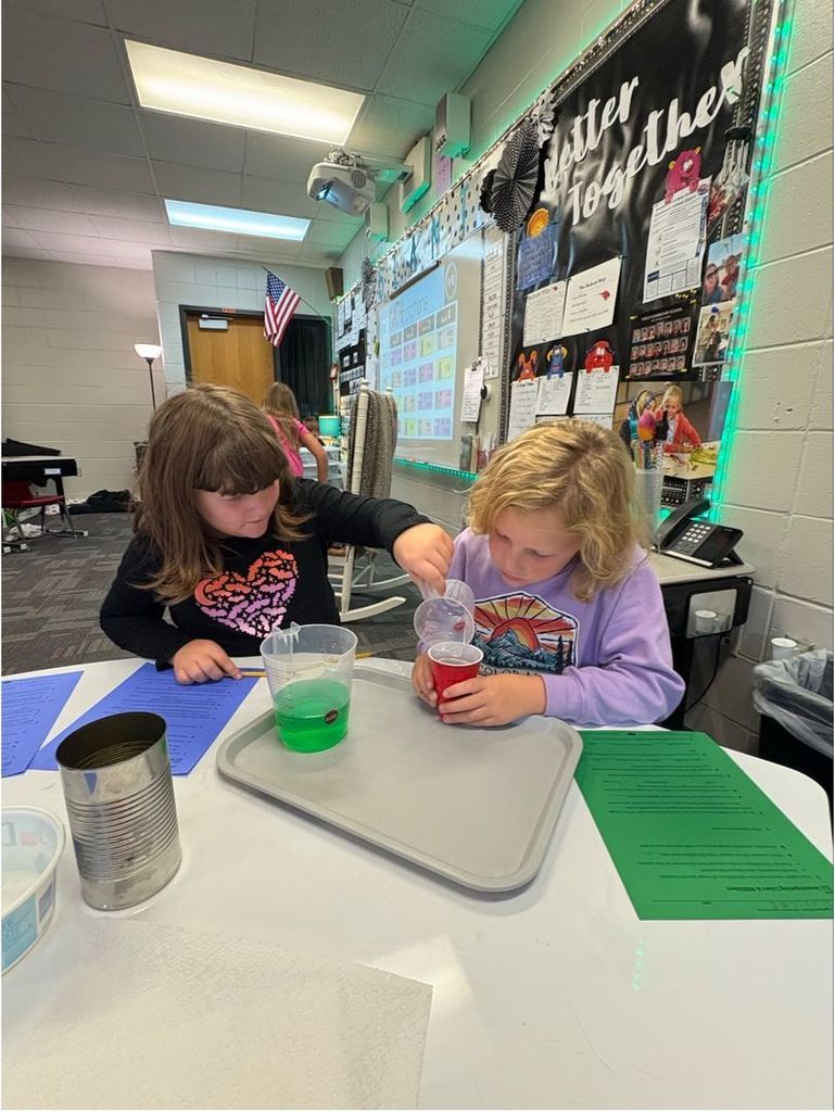 Two students sit side by side measuring green liquid into a red plastic cup as part of a classroom learning activity.