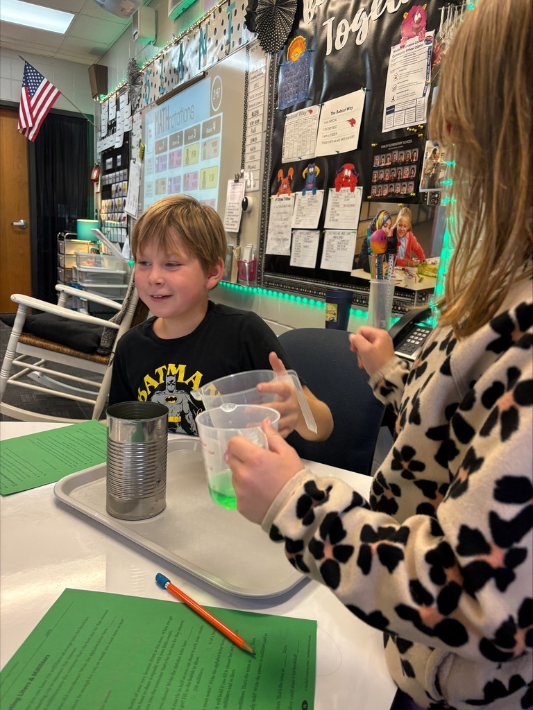 Two elementary students sit at a table in a classroom conducting a hands-on experiment. One student smiles while holding a measuring cup with green liquid, and the other prepares to pour it into a can.