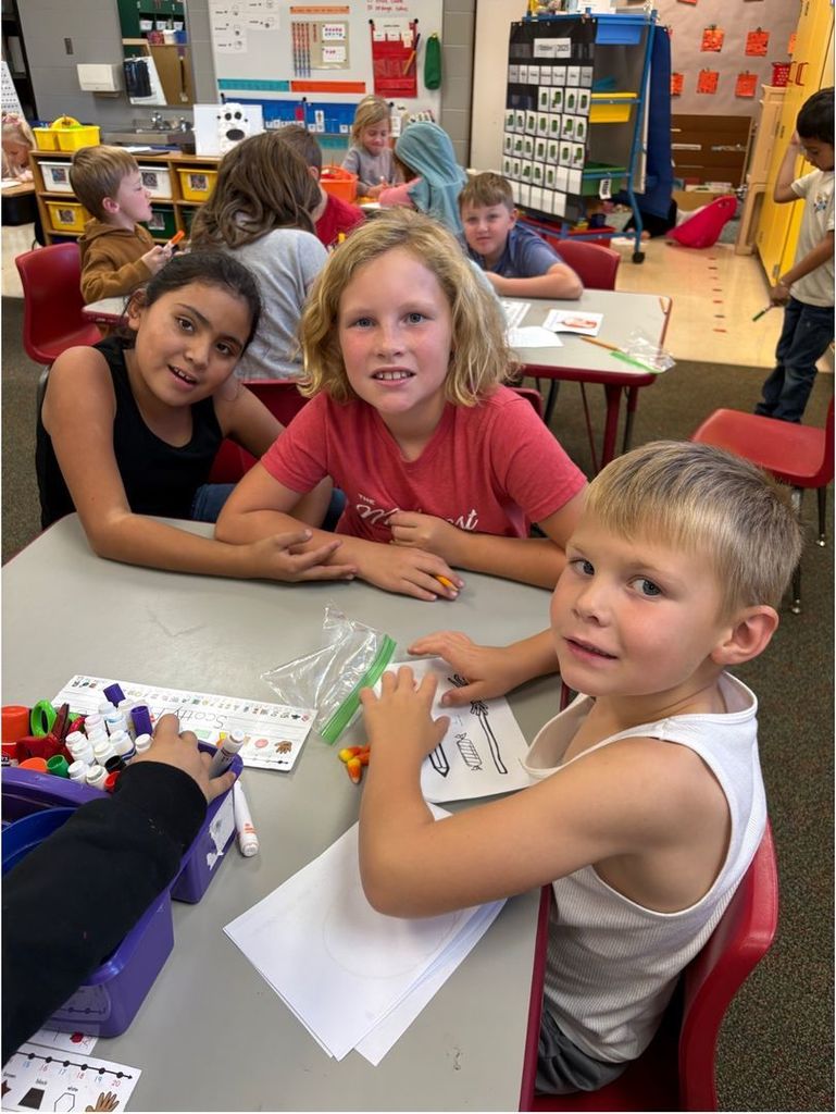 Three students sit together at a table working on a fall-themed classroom activity using candy corn and worksheets. They smile toward the camera.