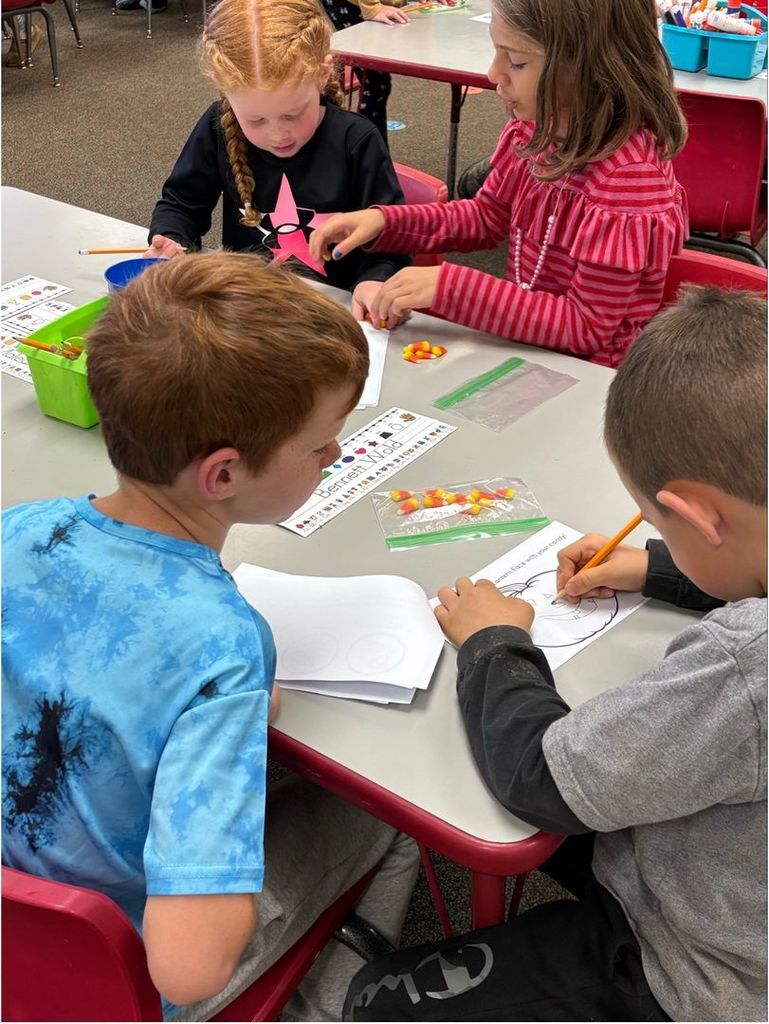 Four elementary students sit together at a classroom table working on a candy corn-themed activity.