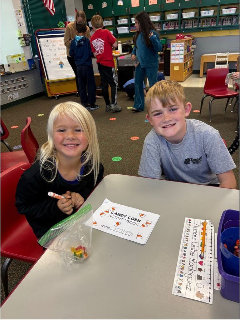 Two smiling students sit at a table with a “Candy Corn Activity Book” and a small bag of candy corn.