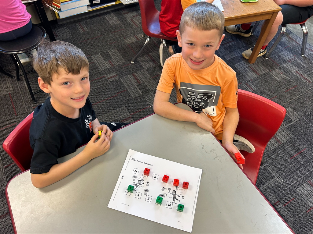 Two students sit at a classroom table smiling while playing a math board game using cubes and dice. A worksheet with numbered squares and illustrations lies in front of them