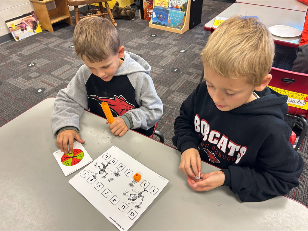 Two students sit together at a desk working on a math activity. One holds a yellow counting block and points to a spinner card, while the other watches attentively.