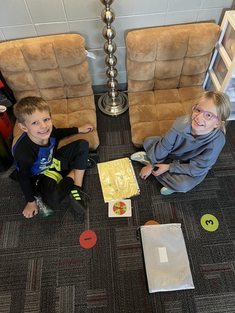 Two students smile while sitting on the classroom floor near tan cushions and a tall lamp. They are playing a math game using worksheets and colorful cards.