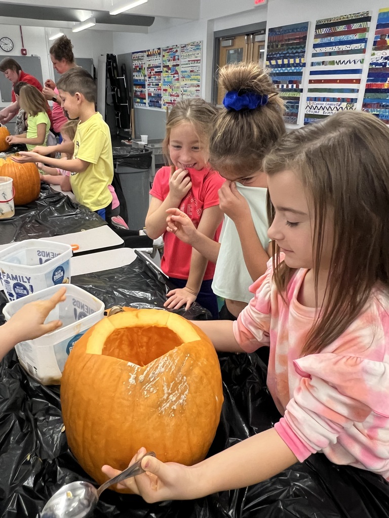 Several students gather around a table cleaning out a large pumpkin. One student uses a spoon to scrape the inside while others smile and laugh, reacting to the messy pumpkin contents.
