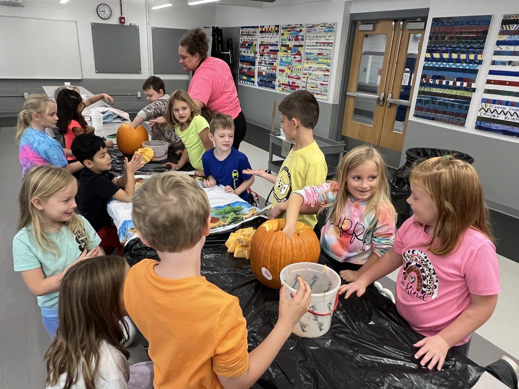 A group of elementary students sit around long tables covered with plastic as they scoop seeds and pulp from pumpkins.