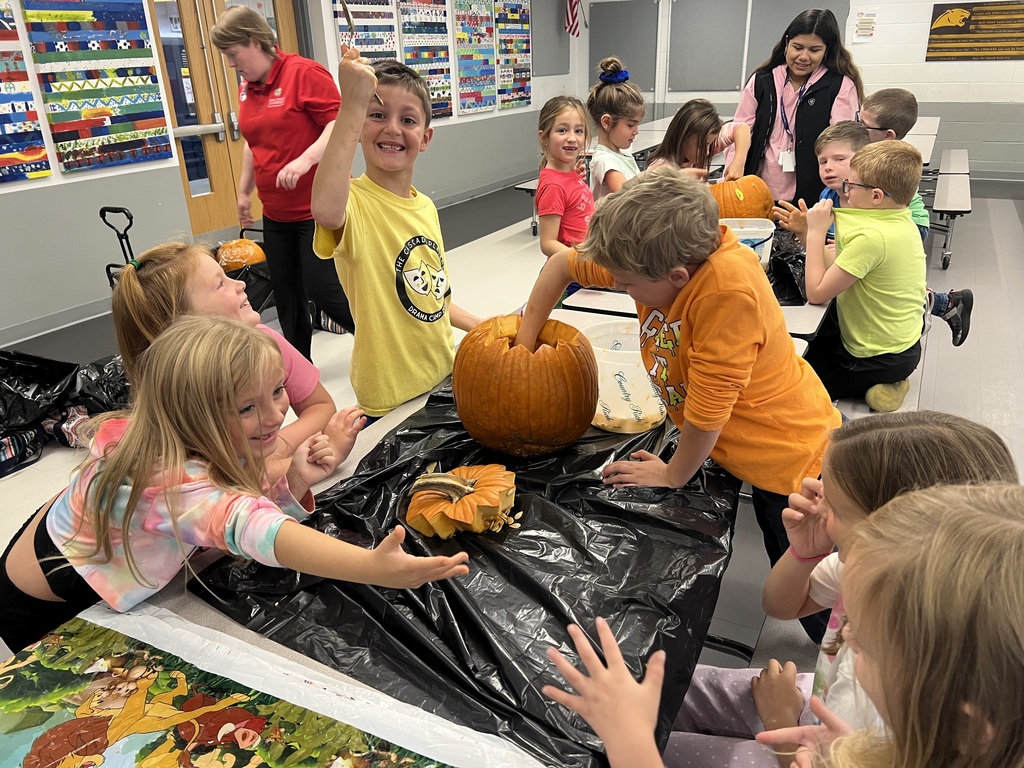 Excited students work together to clean out pumpkins during a classroom activity. One student proudly holds up a spoon, while others reach into the pumpkins and chat with friends.