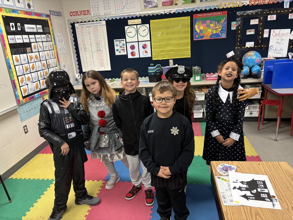 Six students pose in a classroom dressed in various Halloween costumes, including Darth Vader, a clown, a police officer, and Wednesday Addams.