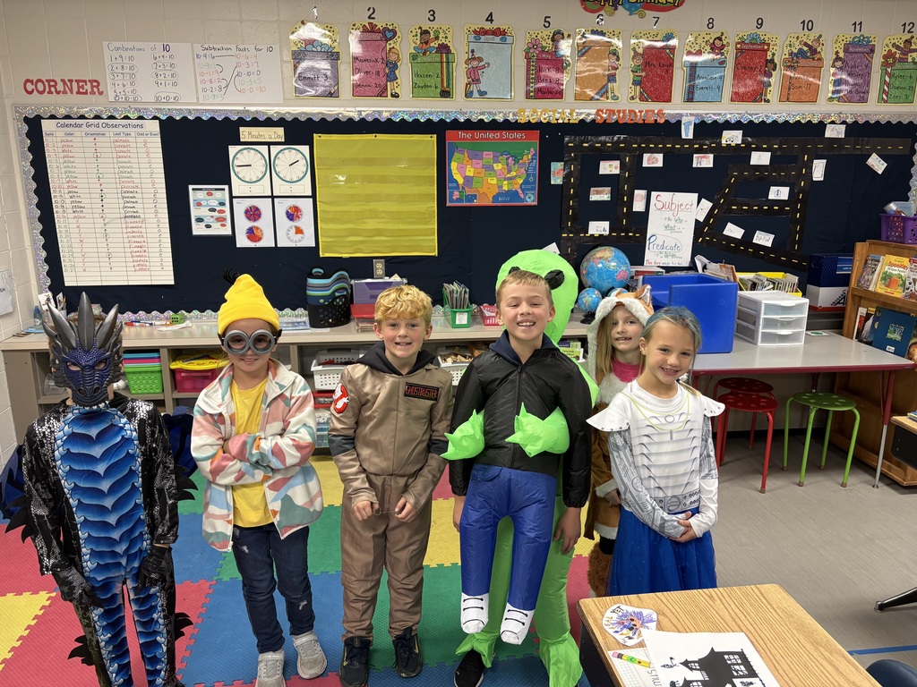 A group of six elementary students stand smiling in a classroom wearing colorful costumes, including a dragon, minion, Ghostbuster, inflatable alien, unicorn, and superhero.