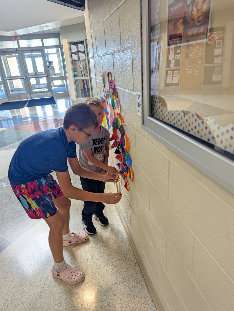 Two students work together in a hallway to add brightly colored paper feathers to a turkey mural on the wall.