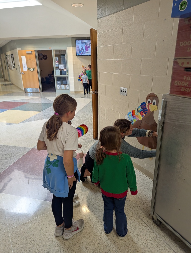 A teacher helps a small group of students attach colorful feathers to a paper turkey on the hallway wall. Two other students watch and wait their turn.