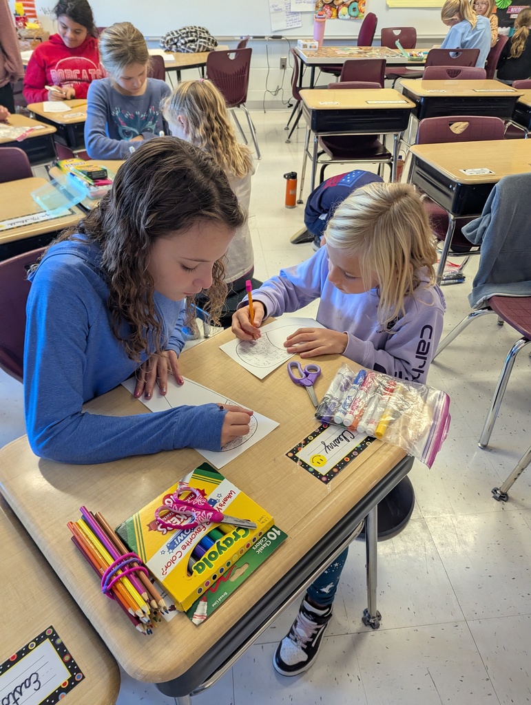 In a classroom, two students sit at a desk coloring paper feathers for a Thanksgiving project.