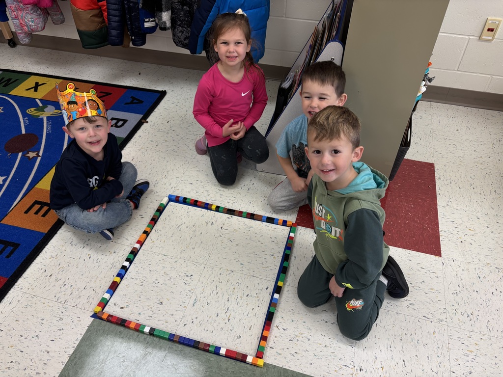 Four young students smile and kneel on the classroom floor beside a large square they built out of colorful plastic cubes.